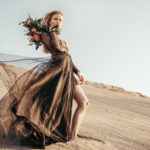 female model posing on beach sand in front of gray blue sky, in a golden dress, exposing her one leg, holding a bouquet of flowers over her shoulder
