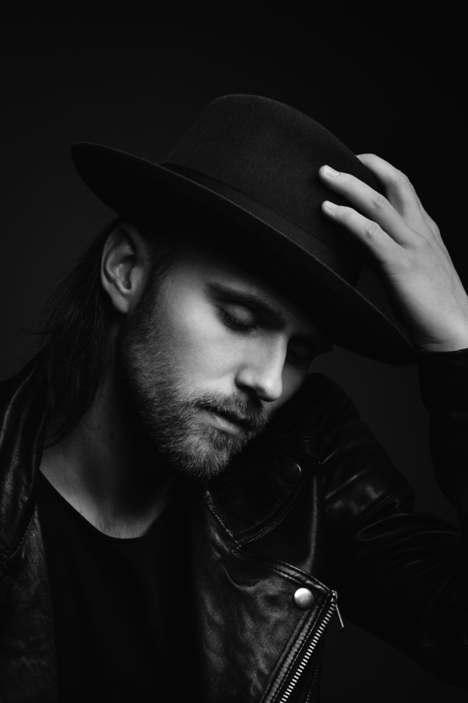 Black and white picture of Lukas Meijer in a hat, holding his hat with one hand and looking away from the camera