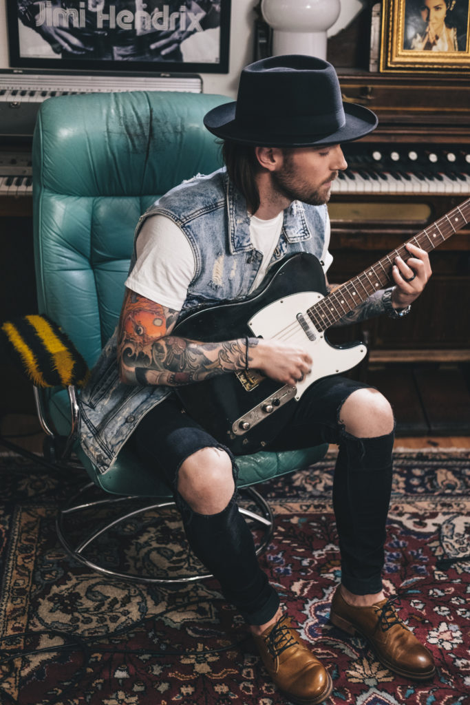 Photo of Lukas Meijer sitting in a blue chair, on a colorful carpet, playing a electric guitar