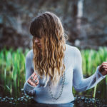 female model posing in blue shirt, looking to the side with her hair covering her face, with grass strands in the background