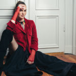 female model posing in a red jacket, red fingerless gloves, sitting against a white door