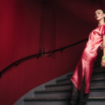 female model posing in pink dress standing on a staircase in front of a red wall